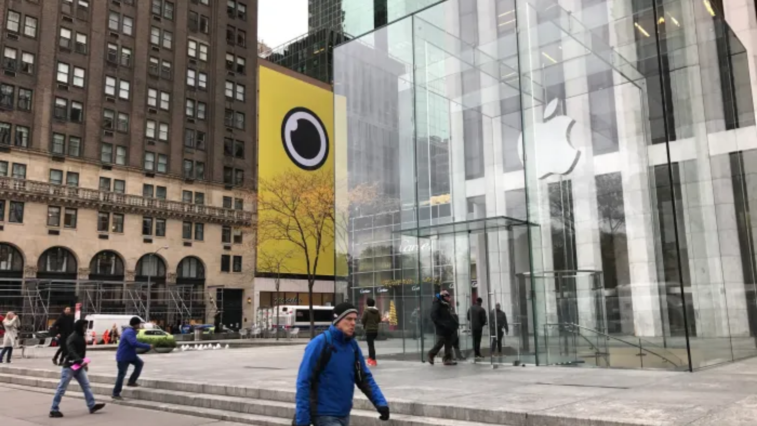 Yellow Spectacles lens-logo building wrap on a Fifth Avenue storefront beside the glass Apple Store cube in Manhattan