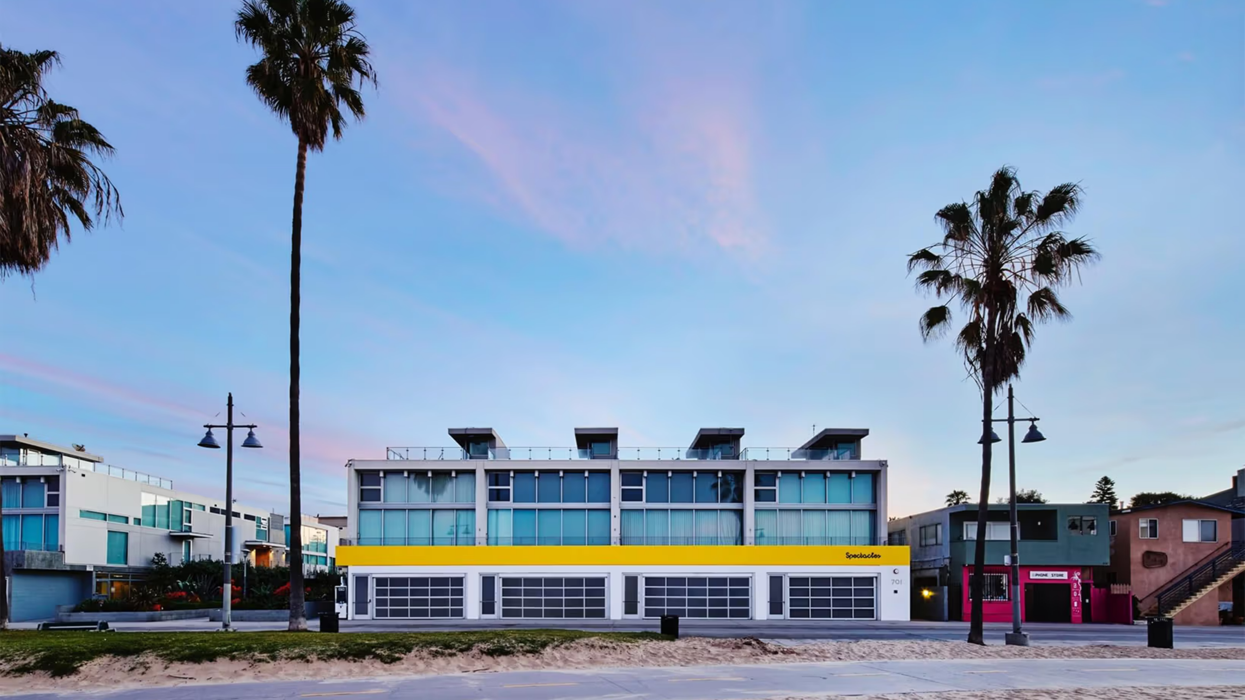 Dusk beachfront building with a bright yellow Spectacles storefront band above roll-up doors, palm trees on either side