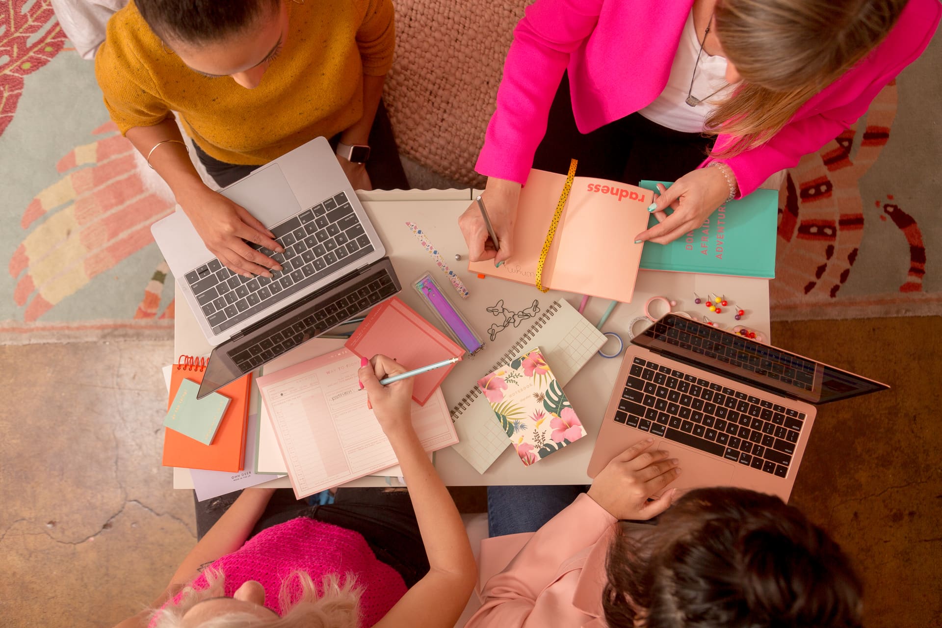 Overhead shot of four people working at a table covered in laptops, pastel notebooks, planners and stationery