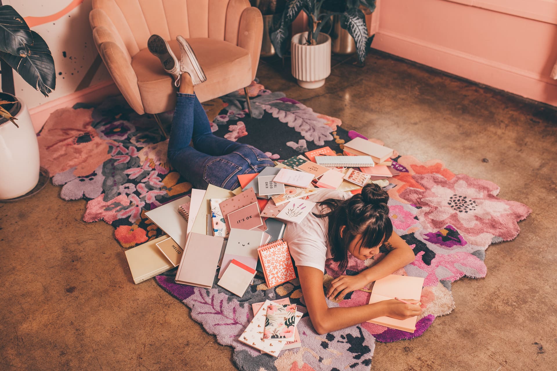 Woman lying on a floral rug writing in a pink notebook, surrounded by stationery, greeting cards and journals