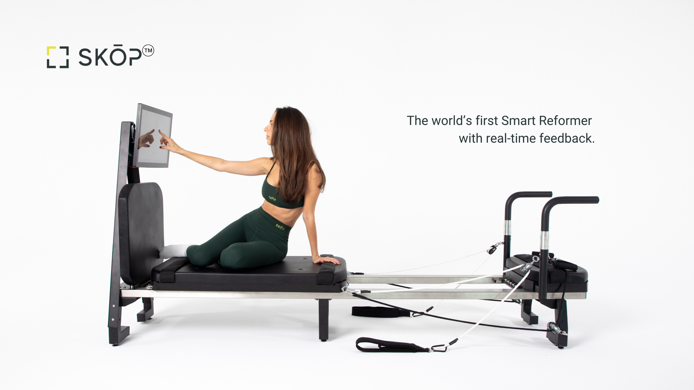 SKOP pilates reformer on white: a person in green activewear sits on the black carriage touching a mounted screen
