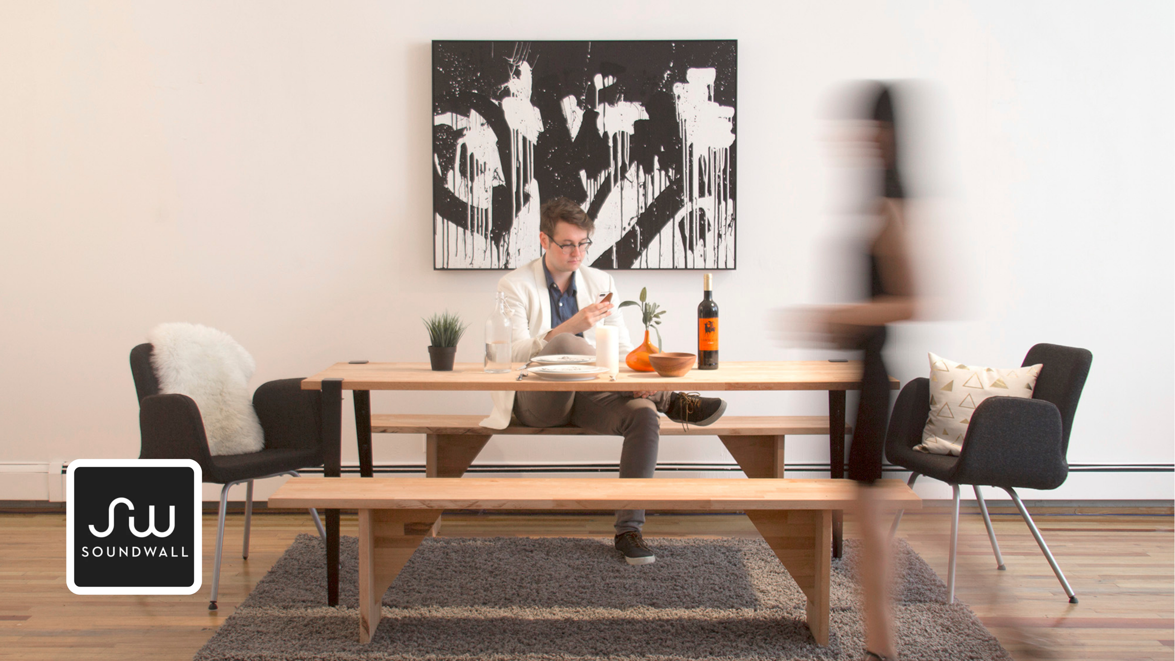 Soundwall card: a black-and-white splatter canvas hangs above a wood table where a person sits with a phone