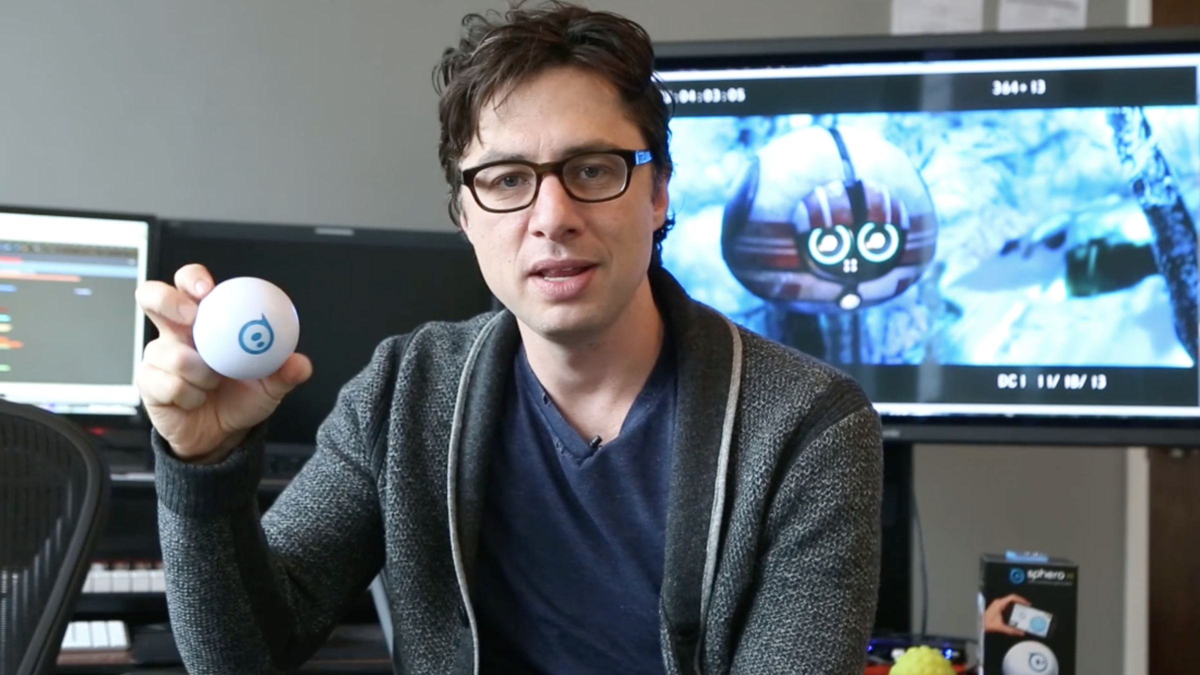 A person in glasses and a gray cardigan holds up a white Sphero ball at a desk, edit monitor glowing behind