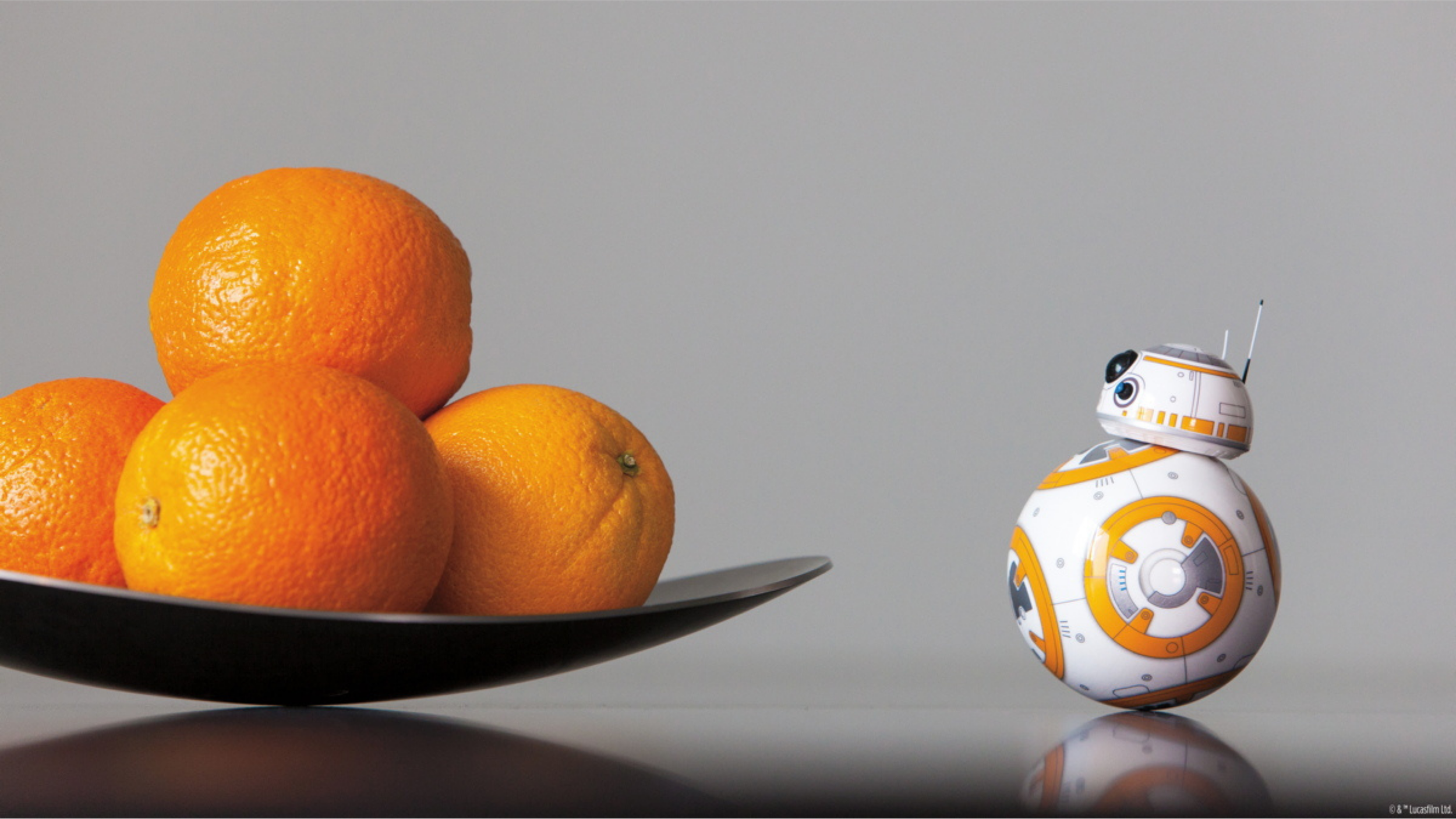 Sphero BB-8 app-enabled droid on a reflective surface beside a black bowl of oranges