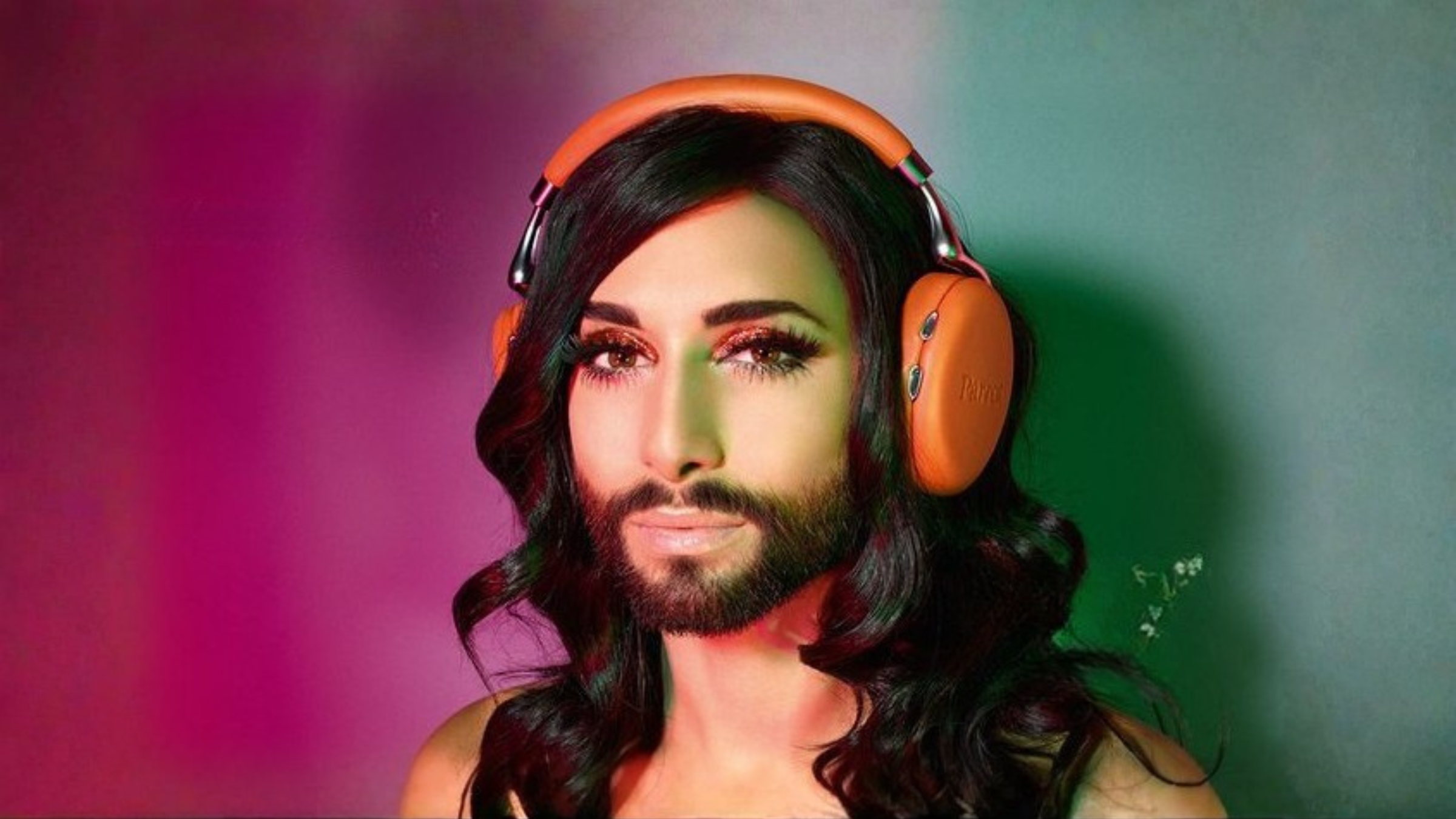 Person with long dark hair and a beard in orange Parrot Zik headphones, lit magenta and green, looking at the camera