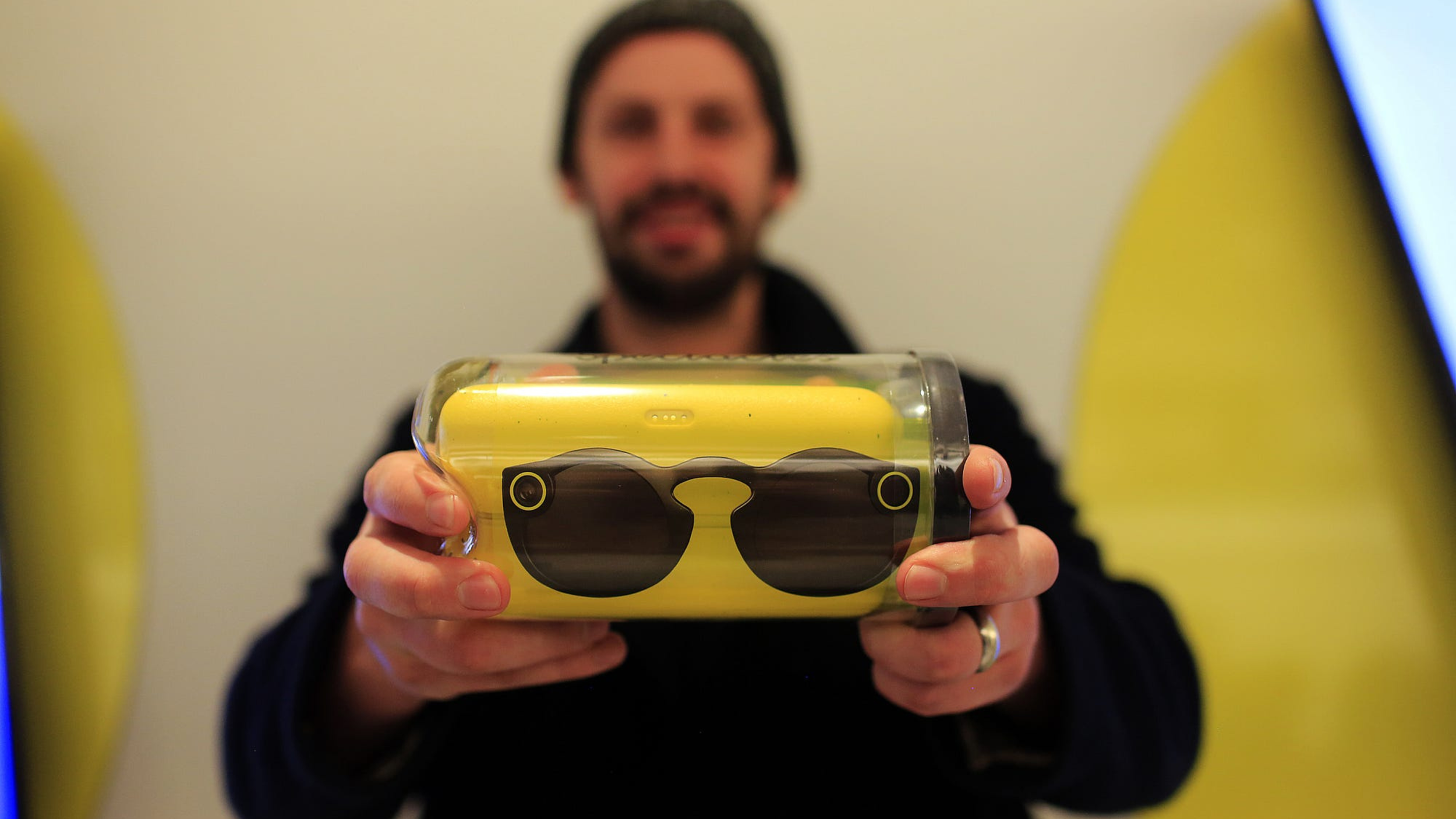 Man in a beanie holds up black Spectacles sealed inside a clear cylindrical package