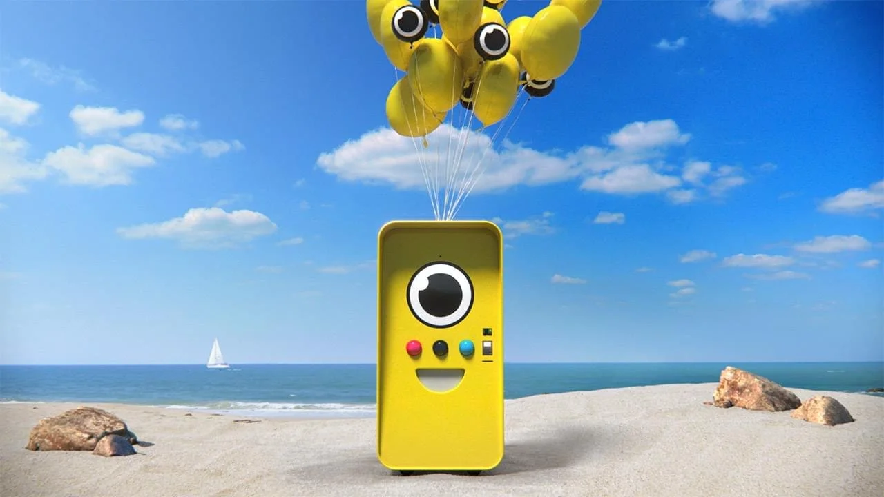 Yellow Snapbot vending machine with a camera-eye face, lifted by yellow balloons on a beach
